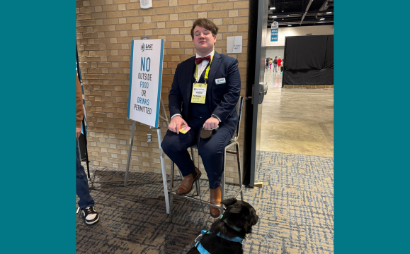 A person in a navy suit with a red bow tie sits on a tall stool beside a sign reading “NO OUTSIDE FOOD OR DRINKS PERMITTED” at an event entrance. The person wears an event badge and holds a phone. A brick wall and open doorway to a large exhibition hall are behind them, with people visible in the distance. In the foreground, a black dog wearing a harness stands on the patterned carpet near the seated person. Provide your feedback on BizChat