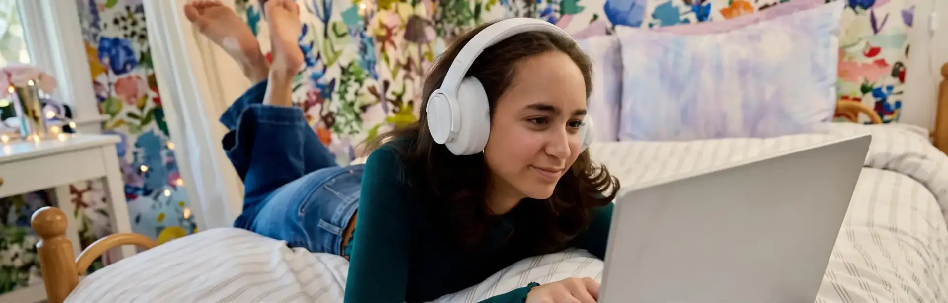 Student laying at her bed wearing a white pair of headphones while using her laptop